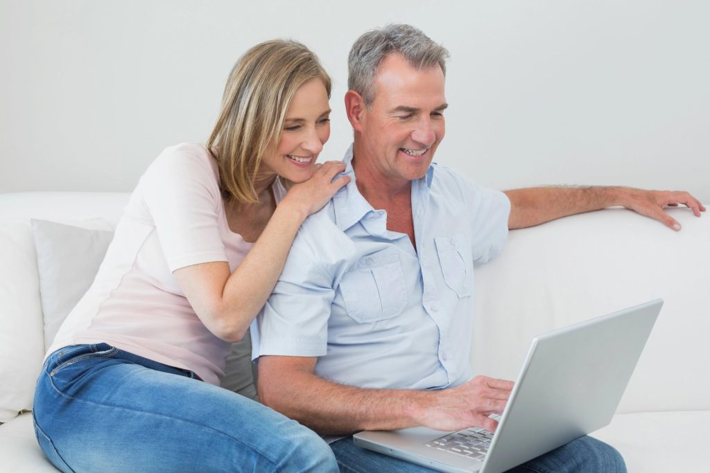 A couple is sitting on a couch, smiling as they look at a laptop together.