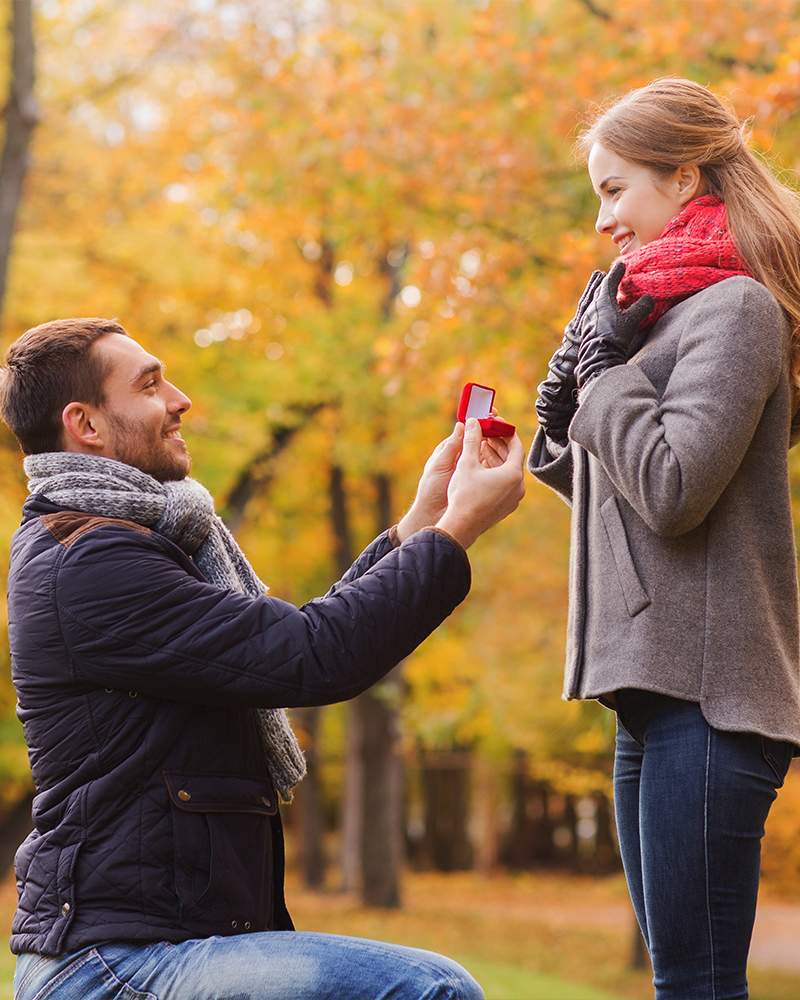 A man is proposing to a woman outdoors, offering a ring. Autumn trees in the background.