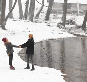 A couple stands in a snowy landscape near a stream, holding hands and smiling at each other.