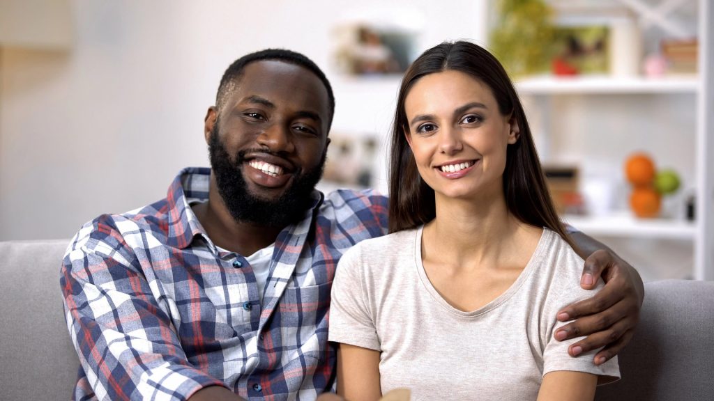 A smiling couple sitting together on a couch in a cozy living room setting.