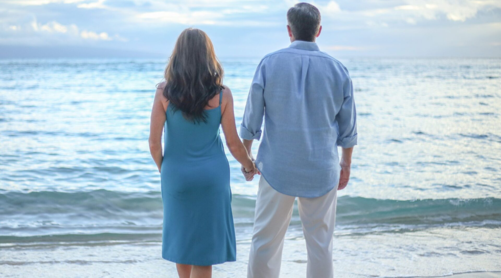 A couple holding hands while standing on a beach, looking out at the ocean.