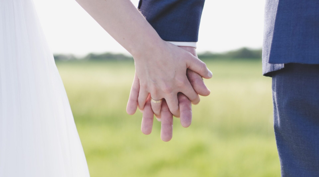 A couple holding hands outdoors, with one wearing a wedding ring.