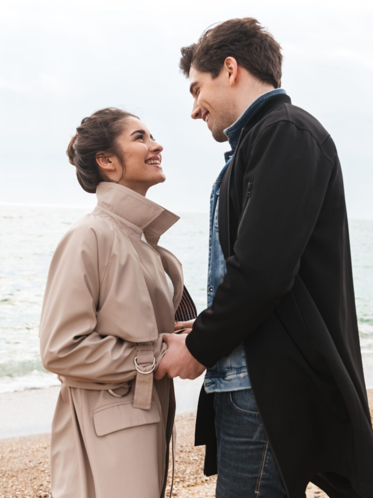 A couple standing on a beach smiling at each other, holding hands, and looking happy.