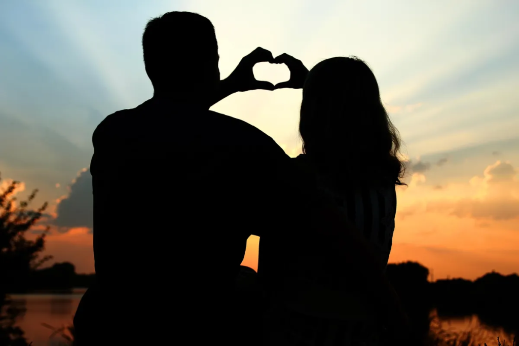 A couple silhouetted against a sunset, forming a heart shape with their hands.