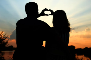 A couple silhouetted against a sunset, forming a heart shape with their hands.