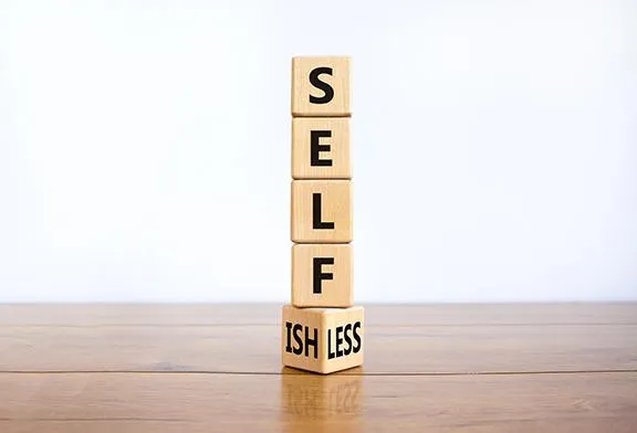 Wooden blocks stacked to spell "SELFISH" and "SELFLESS" on a wooden surface.