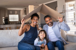 A family of three smiling on a couch, holding a cardboard roof above them, symbolizing a home.