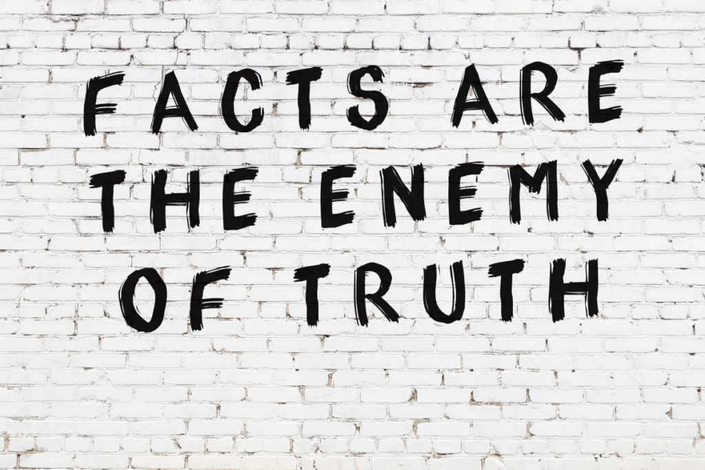 "FACTS ARE THE ENEMY OF TRUTH" is written on a white brick wall.