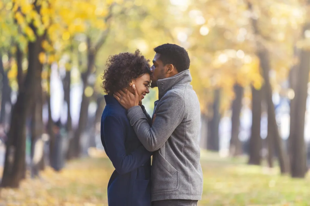 A couple shares an intimate moment in a park with autumn leaves, embracing affectionately.