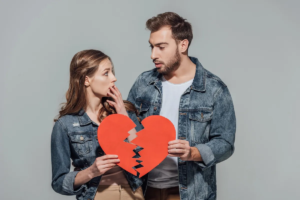 A couple holds a broken paper heart, suggesting relationship issues or a breakup.